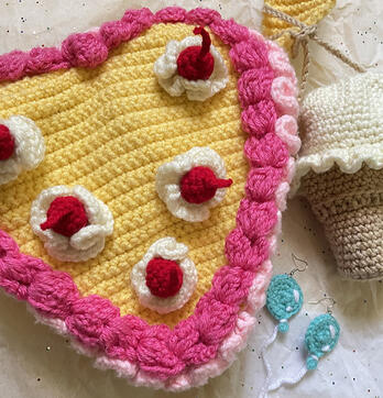 Group of crocheted objects, a yellow heart-shaped cake bag with two layers of pink frosting (one light, one dark), whip cream, and cherries on top, an ice cream cone pouch, and a pair of cyan balloon earrings.
